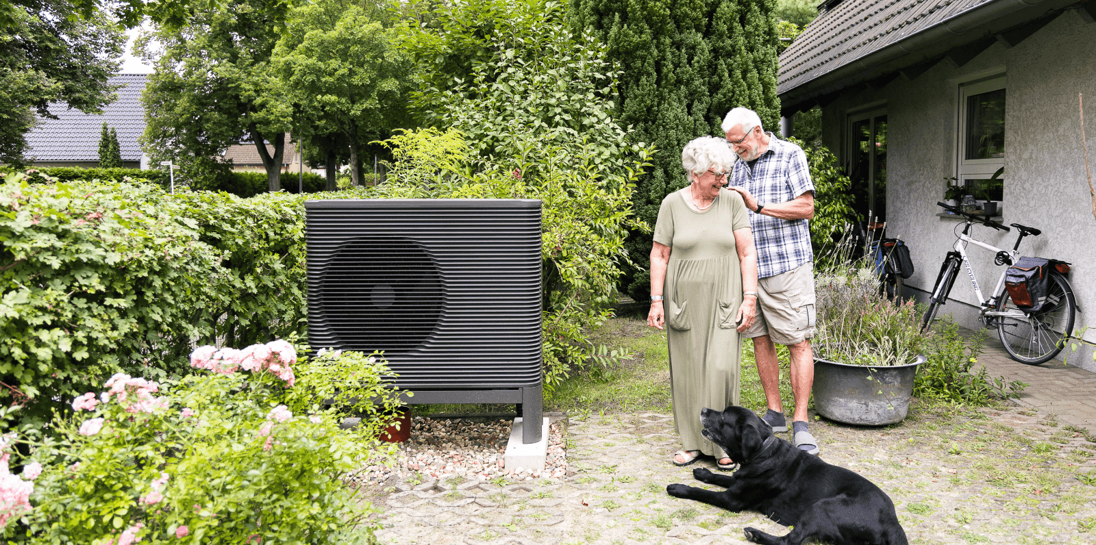 älteres Ehepaar steht mit Hund neben Wärmepumpen Außeneinheit im Garten und lacht